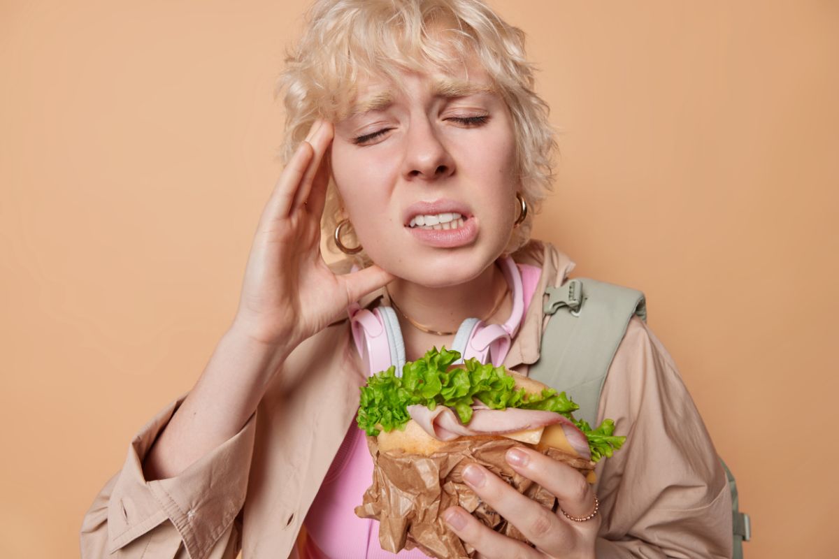 Young woman experiencing sharp tooth pain while biting into a sandwich, a common symptom of dental decay or a cracked tooth.